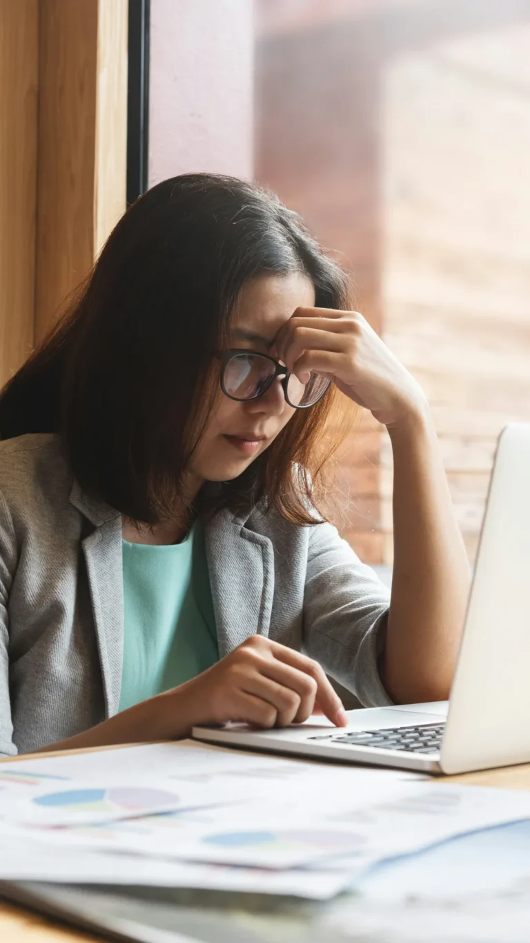 A person working on a laptop with printouts of pie charts and their fingers on their forehead in an expression of some slight distress