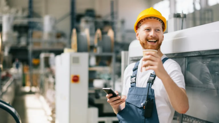 A worker in dungarees and a hardhat smiling and drinking coffee or tea while holding his phone
