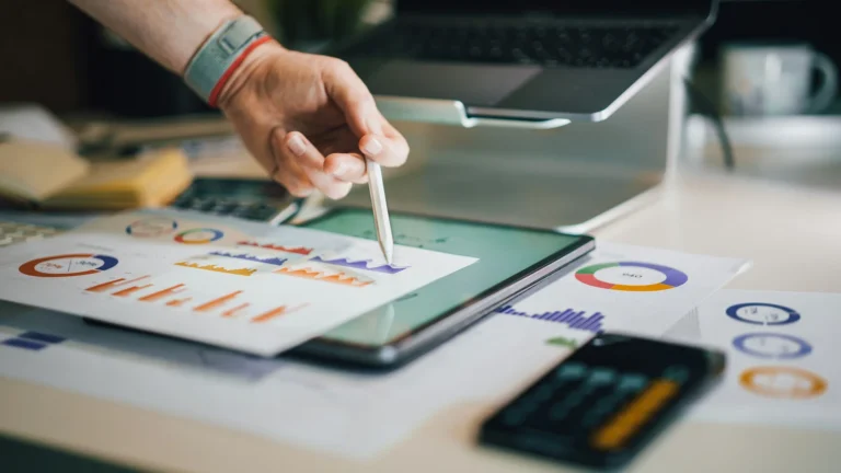 A person with lots of printed donut and bar charts, an a tablet, laptop and calculator on their desk, pointing at one chart with a digital pencil